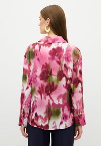 Blusa de manga larga en una tela rosa con estampados florales en tonos de rosa oscuro y verde. Presenta un cuello y puños abotonados. Textura suave.