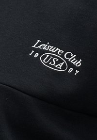 Μαύρο ύφασμα με λευκό κεντημένο κείμενο «Leisure Club 1997» και «USA» μέσα σε οβάλ σχήμα.