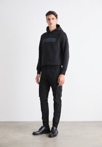 Hanorac negru cu logo frontal, asortat cu pantaloni cargo negri cu buzunare și pantofi negri. Design plat, fără modele sau accente vizibile.