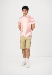 Camicia a maniche corte a righe rosse e bianche, abbinata a pantaloni cargo beige e sneakers bianche, presentata su uno sfondo neutro.