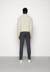 Uomo in piedi di fronte a un muro, che indossa un maglione a collo alto color crema, pantaloni grigi scuri su misura e sneakers bianche.