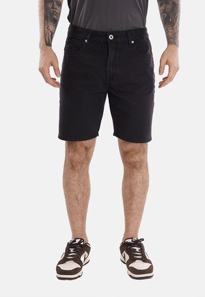 Shorts in denim nero con un design a vita media, tasche frontali e chiusura con bottone. Abbinati a sneakers marroni e bianche.