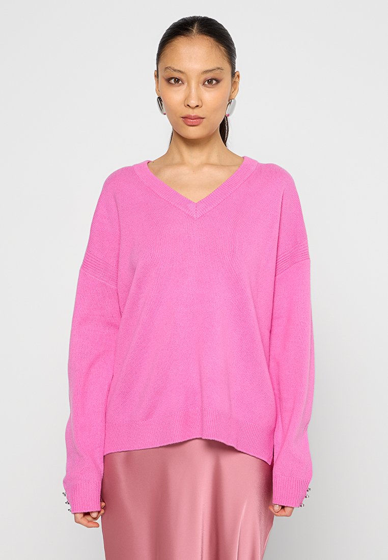 Suéter rosa oversize con escote en V y textura acanalada. Mangas largas con aberturas laterales. La modelo lo lleva sobre una falda de satén rosa.