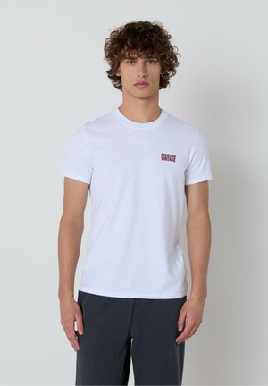 Bela bombažna majica s krožnim izrezom in kratkimi rokavi, z majhnim rdečim logotipom "AMERICAN VINTAGE" na levi strani prsnega koša.