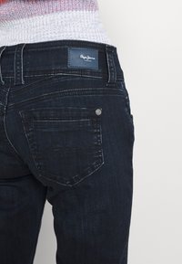 Dark blue denim jeans featuring a straight cut, a leather logo patch on the back waistband, and subtle stitching accents on pockets.