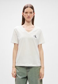 Biela bavlnená tričko s výstrihom do V, krátkymi rukávmi, voľným strihom a čiernym logom "CK" na ľavej strane hrudníka. Mäkká, hladká textura.
