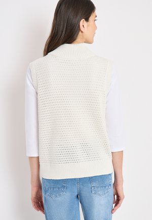 Femme portant un gilet sans manches en tricot crème par-dessus un haut blanc et un jean bleu clair, vue de dos sur un fond uni.