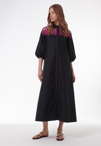 Robe maxi noire avec un col brodé violet, des manches longues bouffantes et une fermeture à boutons sur le devant. Tissu lisse avec une coupe décontractée.