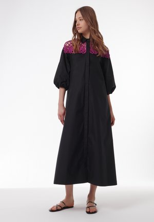 Zwarte maxi jurk met een paarse geborduurde kraag, lange pofmouwen en een knoopsluiting aan de voorkant. Glad materiaal met een ontspannen pasvorm.