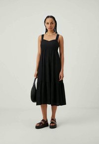 Svart, ärmlös, lager-på-lager klänning i lätt tyg med texturerade detaljer. Modellen har på sig kraftiga sandaler och håller en svart handväska.