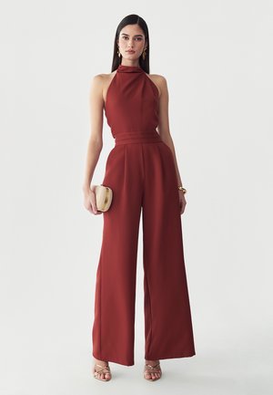 Femme aux longs cheveux foncés portant une combinaison sans manches rouge rouille à col montant, tenant une pochette tressée beige et portant des sandales dorées.