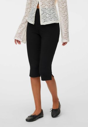 Femme portant un pantalon court noir, des ballerines noires et un haut blanc en dentelle à manches longues avec fermeture boutonnée à l'avant.