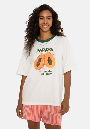 Žena se usmívá, má na sobě bílý oversized tričko s grafikou papáji a textem "PAPAYA mě k tomu přiměla," v kombinaci s oranžovými a růžovými pruhovanými šortkami.