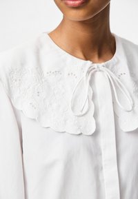Weißes Blusenshirt mit einem gestickten, gewellten Kragen, einer Bindeschließe am Hals und einer glatten Baumwollstruktur.