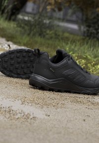 Coppia di scarpe da trail outdoor nere con suole robuste appoggiate su un sentiero di ghiaia con erba sullo sfondo.
