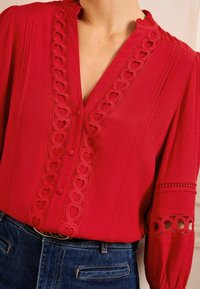 Blusa rossa con bordi in pizzo a forma di cuore, bottone frontale, scollatura arricciata e maniche lunghe con dettagli a taglio. Abbinata a denim.