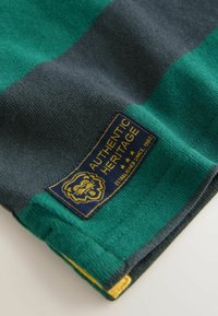 Zelená a námořnická pruhovaná látka s texturovaným povrchem. Obsahuje zlatou nášivku s logem lva, označenou "Autentická dědictví" a žluté akcenty.