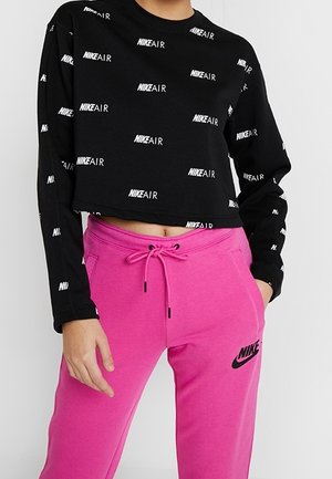 Osoba nosząca czarną krótko przyciętą bluzkę z długim rękawem z białym tekstem "NIKE AIR" oraz jaskrawo różowe spodnie dresowe Nike ze sznurkiem w pasie.
