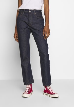 Personne portant un jean bleu foncé à jambes droites, un t-shirt blanc et des baskets montantes rouges, debout devant un fond uni.