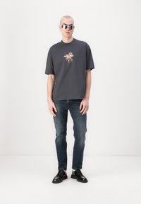 Camiseta gris de algodón con un gráfico floral y texto en el pecho, combinada con jeans azul oscuro y zapatos negros. El modelo lleva gafas de sol reflectantes.