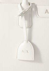 Bolsa de couro branco com textura suave, apresentando uma alça de etiqueta costurada e uma etiqueta retangular estampada com "A/X" em cor suave.