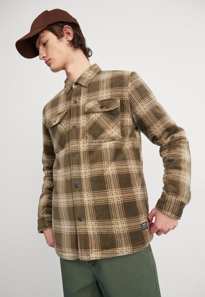 Camisa de franela a cuadros marrón y beige con dos bolsillos en el pecho y botones oscuros. La tela parece suave y texturada. Se lleva con pantalones verdes.