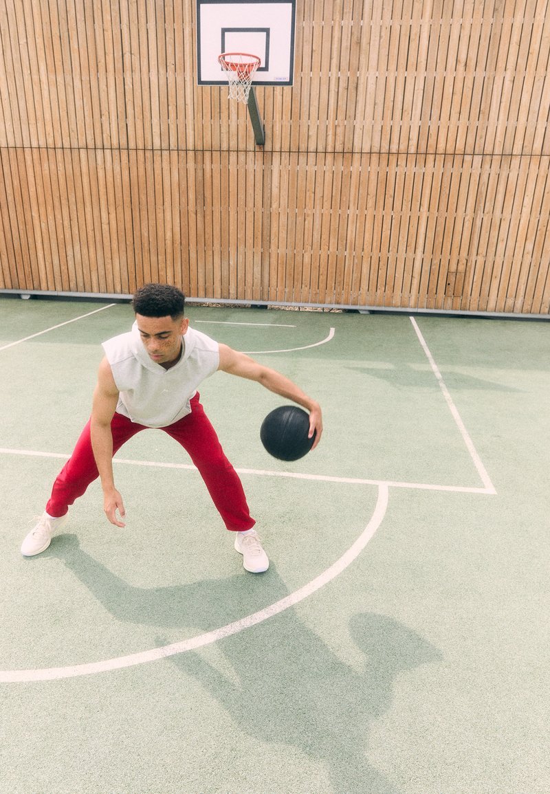Ein junger Mann in einem ärmellosen grauen Shirt und roten Hosen dribbelt einen schwarzen Basketball auf einem grünen Platz mit einer Holzrückwand.