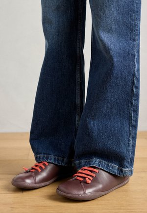 Braune Lederschuhe mit roten Schnürsenkeln, getragen mit weit ausgestellten blauen Jeans, auf einem Holzfußboden präsentiert. Strukturierte Oberfläche, schlichtes Design.