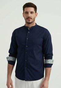 Chemise bleu marine à col mandarin et poignets à motifs. Elle présente des manches longues, un tissu lisse et des détails intérieurs colorés contrastants.