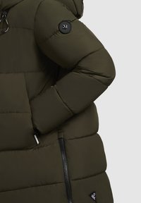 Jachetă pufoasă, de culoare verde măslinie, cu glugă, având un emblemă neagră pe mânecă, o pungă cu fermoar și o textură netedă.