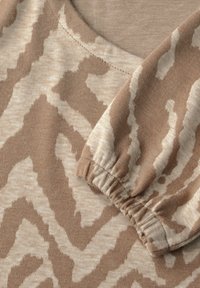 Hellbrauner Stoff mit einem cremefarbenen abstrakten Zickzack-Muster. Verfügt über einen runden Halsausschnitt und elastische Bündchen an den Ärmeln. Weiche Textur.