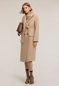 Cappotto di lana beige con design doppiopetto, ampio colletto e tasche laterali. Abbinato a una sciarpa coordinata e chic scarpe col tacco color cammello.