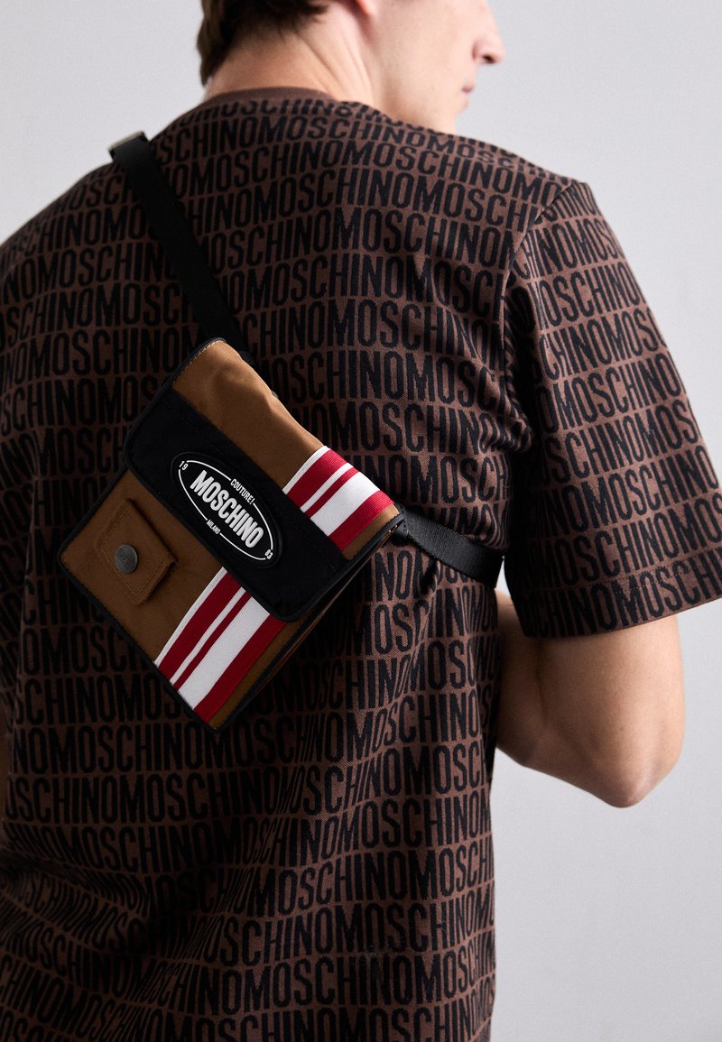 Sac d'épaule à motifs marron avec un panneau noir, présentant des rayures rouges et blanches et un logo. Le porteur porte un t-shirt marron imprimé.