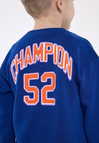 Modrá mikina má na zádech vyšitý text "CHAMPION" a "52" v oranžové a bílé barvě. Měkká tkanina s kulatým výstřihem a dlouhými rukávy.