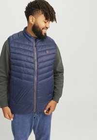 Dunkelblau gesteppte Pufferweste mit hohem Kragen, Reißverschluss und Logopatch, getragen über einem grauen Langarmshirt, kombiniert mit Jeans.