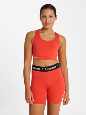 Kvinde iført rød sports-bh og matchende højtaljede shorts med sort hummel-mærket elastik i taljen, stående mod en hvid baggrund.