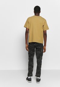 Man met de rug naar de camera gekeerd, gekleed in een mosterdgele korte mouwen shirt, zwarte camouflage cargobroek, witte sokken en zwarte sneakers.