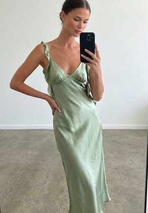 Ženska, ki si v ogledalu posname selfi, oblečena v svetlo zeleno satenasto obleko z tankimi naramnicami in detajli z volančki na enostavni podlagi.