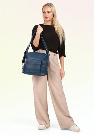 Blonde vrouw in zwarte top en beige wijde broek houdt een grote blauwe leren handtas over de schouder, staand op een effen achtergrond.