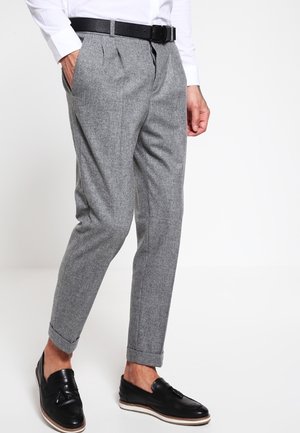 Mann trägt graue konisch geschnittene Anzughose mit umgeschlagenen Säumen, schwarzen Gürtel, weißes Hemd und schwarze Leder-Tasselloafer, steht mit einer Hand in der Tasche.