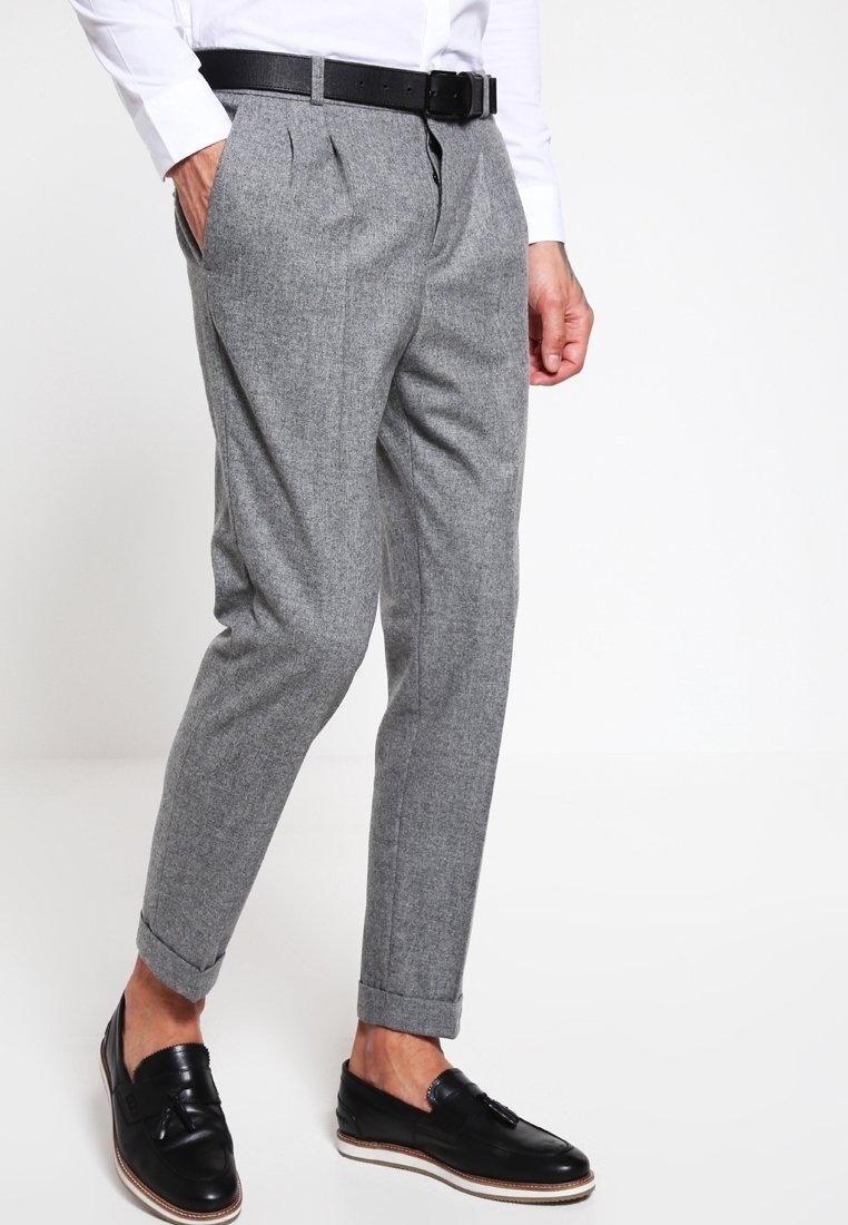 Homme portant un pantalon habillé gris à coupe fuselée avec ourlets retroussés, ceinture noire, chemise blanche et mocassins en cuir noirs à pampilles, debout avec une main dans la poche.