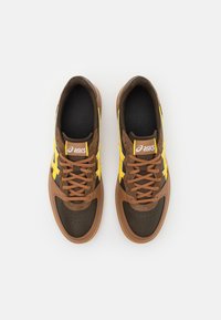 Chaussures de sport marron avec des accents jaunes et blancs. Elles présentent un dessus en cuir lisse et une semelle texturée. Les lacets correspondent à la couleur du dessus.