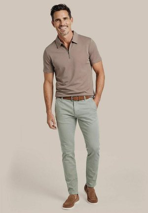 Lachende man staand met één hand in de zak, met een bruine polo met korte mouwen, lichtgrijze broek, bruine riem en bruine instapschoenen.