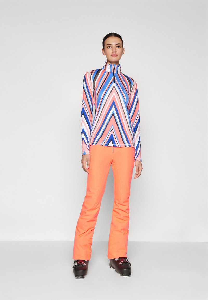 Bunt gestreiftes Longsleeve-Oberteil mit hohem Kragen und eng anliegenden orangen Skihosen. Schwarze Skistiefel vervollständigen das Outfit.