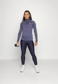 Temno modra športna majica z dolgimi rokavi in polovičnim zadrgom; kombinirana z bleščečimi temno modrimi pajkicami. Modelka drži steklenico z vodo in nosi bele športne copate.