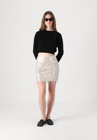 Svart cropped genser, metallisk frynset mini-skjørt og svarte slip-on sko. Modell iført solbriller, står mot en nøytral bakgrunn.