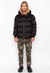 Schwarze Steppjacke mit Kapuze, vorne mit Reißverschluss und elastischen Bündchen. Kombination mit Camouflage-Cargohosen und schwarzen Schuhen.