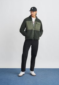 Groene fleecejas met een ontwerp van een effen paneel, ritssluiting en opgestikte zak. Draag met zwarte broek en witte sneakers op een blauwe vloer.