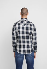 Chemise à manches longues à carreaux avec des carrés bleu marine, blanc et gris. Elle possède un col pointu et un ourlet arrondi. Adaptée à une tenue décontractée.