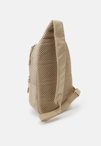 Beige ryggsäck med en texturerad meshnätpanel, rektangulär form, justerbar rem och en blixtlåsficka på sidan.